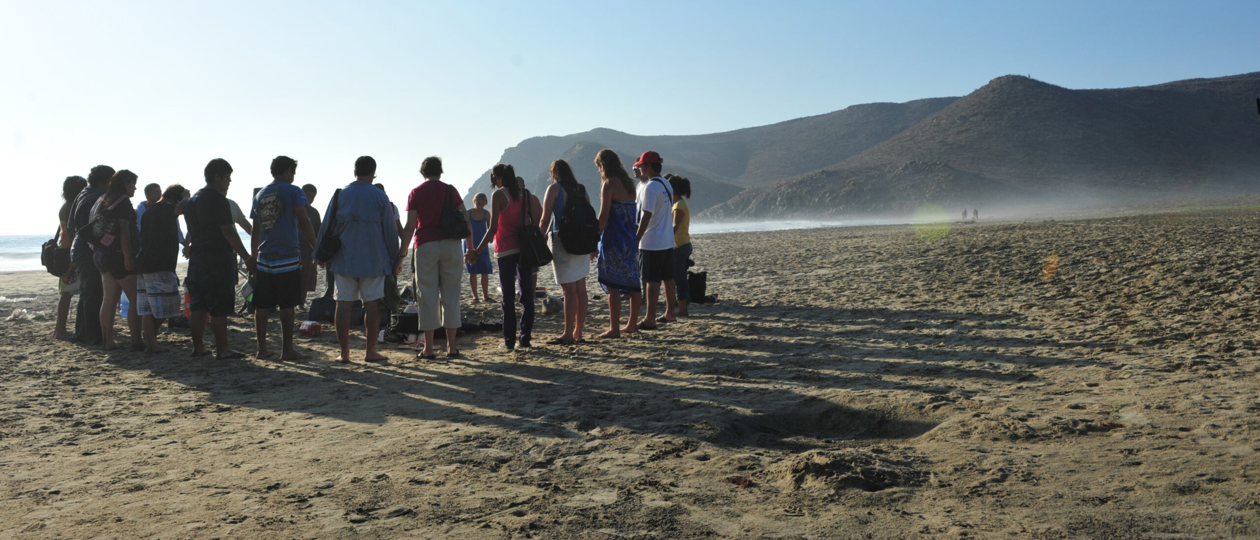 A group stands together praying while holding hands on a beach.